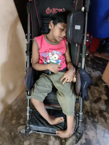 Photo of a small boy from India, aged about 7, sleeping in his wheelchair. He is wearing a pink Winnie the Pooh T shirt and khaki trousers. His feet are bent, so they curl in heavily, rather than going out in front of him.
