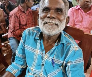 A smiling, older Indian man in a blue checked shirt
