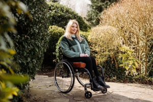 Jade Reynolds: a lady with long blonde hair, sat in a wheelchair, smiling. She is outdoors in a garden, surrounded by trees and bushes.