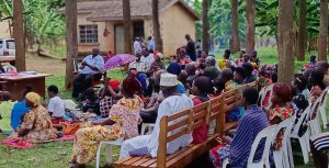 A large group of Ugandan mean and women at a meeting, sitting outdoors on chairs/blankets, wearing colourful clothing, with some in hats/scarves.