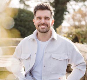 John Reynolds: A smiling man with short brown hair and a short beard and moustache. He is stood outdoors in a garden.