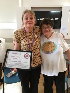 Two ladies side by side, one is holding a framed certificate that reads ‘Luke 5 Award for Christian Disability Inclusion’ and has her arm round the other lady who is wearing a T shirt that reads ‘Rich Tea Community, Fun Faith Friendship’