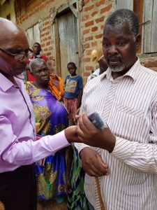 A Ugandan man (Nathan) in a white striped shirt is handed an audio Bible because he is blind