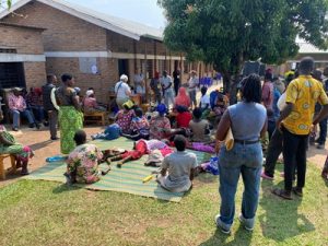 A group of local Rwandan people waiting outside a school building. Many of the people are disabled and are lying on mats.