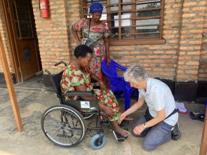 A Rwandan lady sitting in a wheelchair outside a building, with a British lady in a grey T shirt examining how the wheelchair is fitted around the lady's feet.