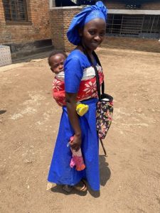 A young Rwandan lady wearing a blue dress and matching blue hat, carrying a baby on her back, strapped to her with a bright red woven shawl.