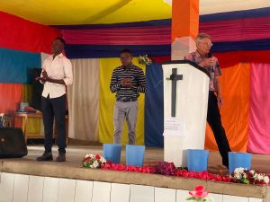 A Rwandan church sanctuary with colourful cloth draped around the altar area. Three men are standing at the front speaking.