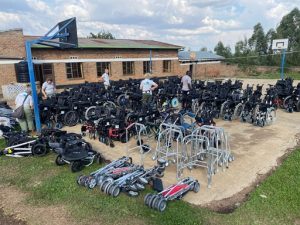 Lots of wheelchairs and mobility aids like zimmer frames laid out in a yard by a building, with people in grey T shirts examining the equipment.