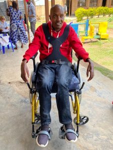 A Rwandan man (Moses) in a red sports jacket, sat in a yellow wheelchair, smiling.