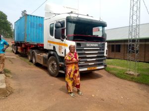 Rwandan lady in a yellow and red dress and headscarf, stood in front of a huge lorry with a shipping container on the back 