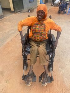 Rwandan man in an orange jacket with a hood up sat in a wheelchair, smiling broadly.