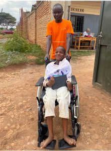 Rwandan boy (Theogene) in a white shirt and trousers sat in wheelchair, being pushed by a friend in an orange T shirt.