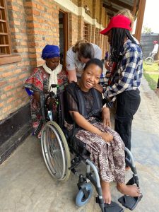 A Rwandan lady (Yvonne) with Cerebral Palsy, sat in a wheelchair, smiling. Behind her, a UK volunteer adjusts her chair, while two local women watch and support.