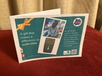 Photo of a hard copy printed gift card on a dark green background, with text on the left that says: 'A gift that makes a difference: an Audio Bible’ To the right are 2 photos: a close-up photo of a solar-powered audio Bible (which looks like a small mobile phone); a smiling young Rwandan man holding an Audio Bible up in the air, as he sits in a wheelchair. Below the images is text saying: 'My gift to you supplies an Audio Bible so a disabled person hears the Word of life in their own language - like this young man in Rwanda. There are logos for Through the Roof www.throughtheroof.org and their project Wheels for the World. There is also a QR code linking to their International Missions page at https://throughtheroof.org/international-mission/