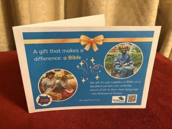 Photo of a printed gift card on a bright blue background, with text on the left that says: 'A gift that makes a difference: a Bible’ Below this is a photo of two female UK volunteers , unpacking Bibles ready to distribute. On the right is an image of a smiling young man from Kenya in a wheelchair; he wears a blue shirt and is holding a paper Bible. Below the image is text saying: 'My gift to you supplies a Bible so a disabled person can read the Word of life in their own language - like Manase in Kenya. There are logos for Through the Roof www.throughtheroof.org and their project Wheels for the World. There is also a QR code linking to their International Missions page at https://throughtheroof.org/international-mission/