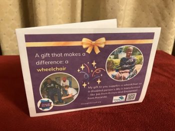 Photo of a printed gift card on a purple background, with text on the left that says: 'A gift that makes a difference: a wheelchair' Below this is a photo of a man in Rwanda being fitted with a wheelchair. On the right is an image of a smiling young man from Kenya in a wheelchair; he has no arms or legs. Below the image is text saying: 'My gift to you supplies a wheelchair so a disabled person's life is transformed - like Job in Kenya and Emmanuel in Rwanda'. showing two people receiving wheelchairs as gifts in Kenya and Rwanda. There are logos for Through the Roof www.throughtheroof.org and their project Wheels for the World. There is also a QR code linking to their International Missions page at https://throughtheroof.org/international-mission/