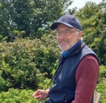 A smiling man with a grey beard, wearing glasses and a dark blue based-ball cap and a blue body-warmer. He is sat on a bench in a garden with trees and bushes behind him. 