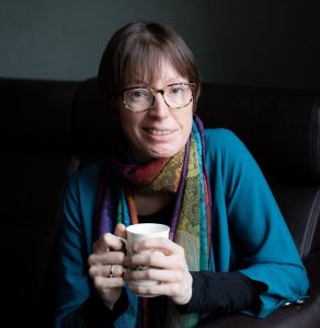 Smiling woman wearing glasses (Emily Owen) who is wearing a turquoise shirt and a colourful scarf, and holding a coffee cup