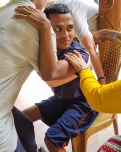 A young man age 28 in navy blue shorts and T shirt, being lowered into a wicker chair by two volunteers.