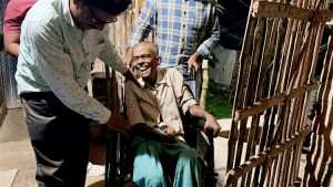 A smiling elderly Indian man, sat in a wheelchair, being pushed by another man, as a further volunteer checks he is comfortable.