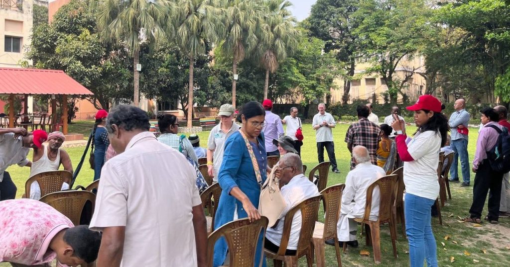 A large group of people in a park in India, outside a block of buildings. There are rows of chairs with people sat, standing, or walking around - men and weomen, old and young, including a man and woman from the UK in grey T shirts.