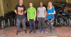 A group photo of 4 UK volunteers in India – one man and three women – stood in front of rows of wheelchairs, lined up for distribution.