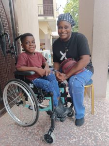 A Nigerian boy and his Mum sat in their kitchen. The boy is aged about 10 and is sat in a wheelchair, smiling broadly.