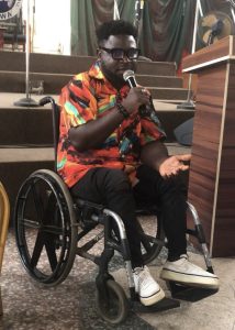 A Nigerian man sat in a wheelchair, holding a microphone, wearing a colourful shirt