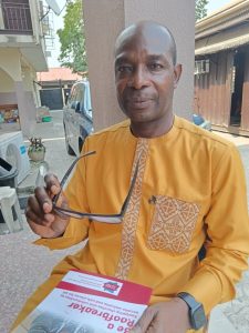A Nigerian man in an orange collarless shirt, holding a pair of glasses and a booklet called 'Be a Roofbreaker'