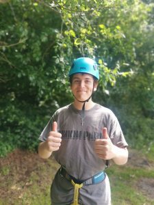 A young man with a blue crash helmet in front of a tree, smiling and holding his hands in a double 'thumbs up'.