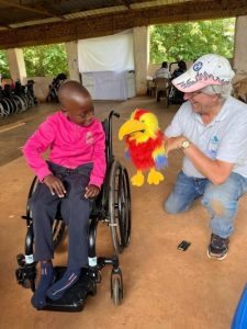 A man with white hair and a white cap showing a red and yellow puppet to a smiling Ugandan child in a wheelchair.