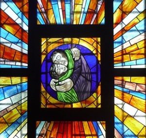 A stained glass window showing two people hugging, platonically, with rays of sunshine around them.