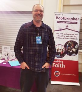 A smiling middle-aged man in a check shirt, stood next to a banner that reads 'Become a Roofbreaker: Enabling faith... Transforming lives through Jesus with disabled people.'