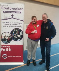 Two men with a arm around each others' shoulders, stood by a banner that reads 'Become a Roofbreaker ... Enabling Faith ... Transforming lives through Jesus with disabled people'.
