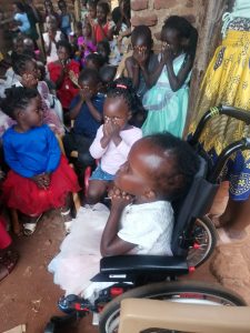 Five year old Ugandan girl in a wheelchair, praying with eyes closed, hands together, as other children pray behind her.