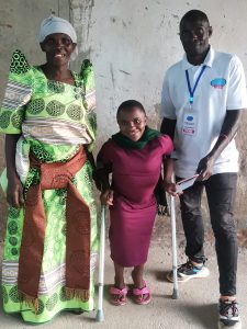 Young Ugandan lady in a purple dress, walking with crutches, stood with a Ugandan male volunteer and a female family member. 