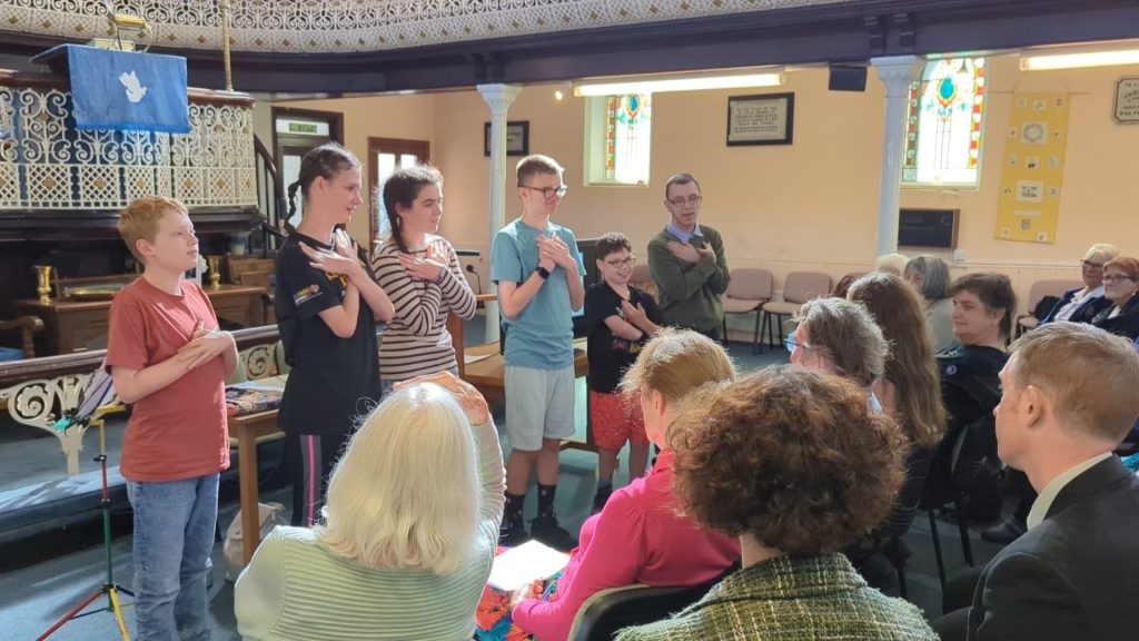 Six young people standing at the front of a church, doing Makaton signs as they sing to a group of older people, sat facing them.