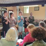 Six young people standing at the front of a church, doing Makaton signs as they sing to a group of older people, sat facing them.