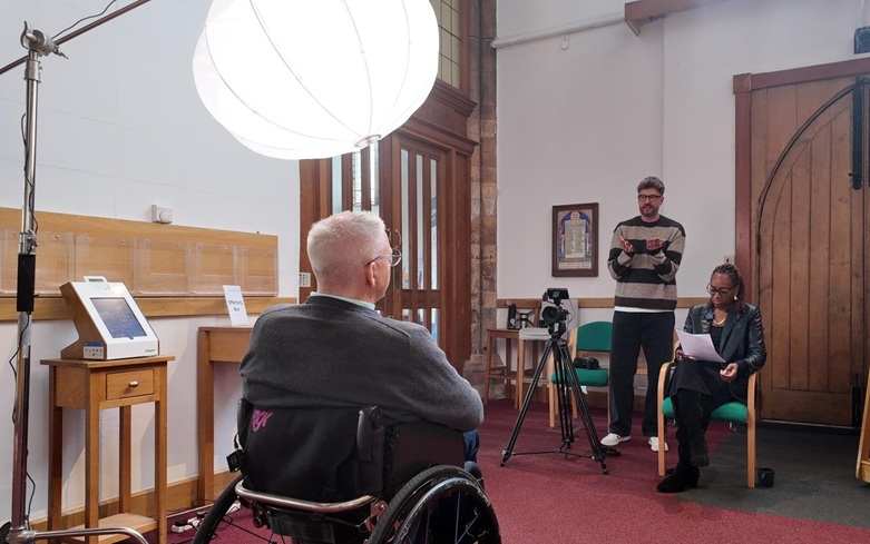 Video filming in a church: a woman in a wooden chair interviews a man in a wheelchair, while a cameraman records it