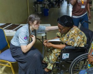 A woman offers prayer and comfort to another woman seated in a wheelchair holding a Bible, indoors.