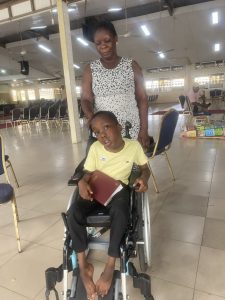 A Ghanaian woman stands behind a boy in a wheelchair who is holding a Bible, inside a large hall lined with rows of empty chairs.