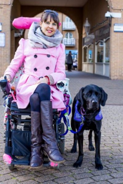 Sally is sat in her pink powered wheelchair, wearing a pink coat, next to her assistance dog, Ethan