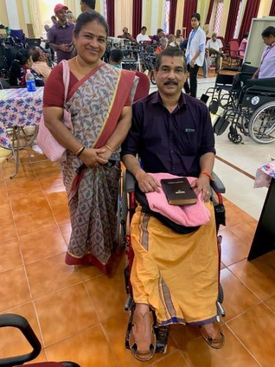 An Indian couple -- a man is sat in his new wheelchair, with a Bible on his lap, and a woman is standing next to him