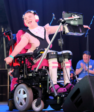 Becky is sat in her wheelchair on the stage preaching at Greenbelt Festival. Her arms are outstretched, and she's using her eyegaze device to speak.
