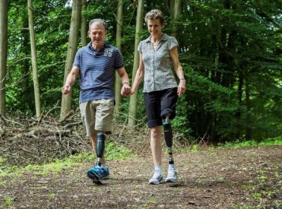 Chris and Denise walking along a wooden path. They're holding hands and smiling. Both of them have a left prosthetic leg, beginning at around the knee.