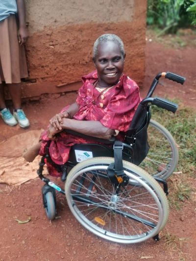 An elderly Ugandan lady, smiling, sat in a wheelchair,  by a mud hut.