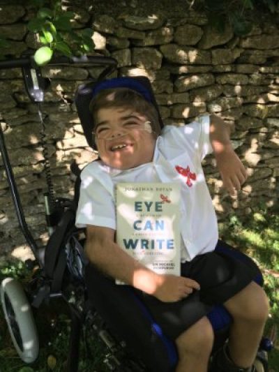 Jonathan is sat in his wheelchair, holding a copy of his book 'Eye can write'. He's grinning and looking towards the camera.