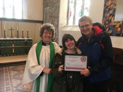 Elana holds the Luke 5 certificate, with Julie the vicar, and Steve on either side of her. They're all standing near the altar in her church.