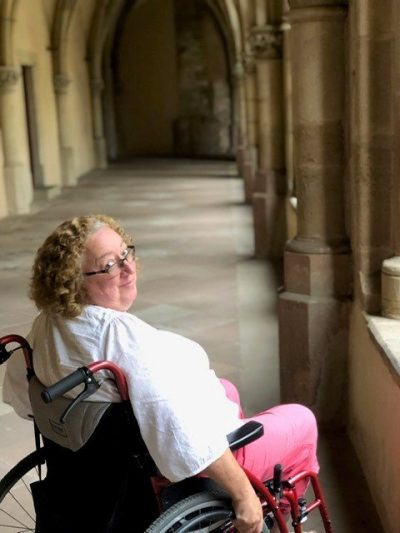 Sarah sits in her wheelchair in a corridor in an abbey or church. She's looking back over her shoulder at the camera.