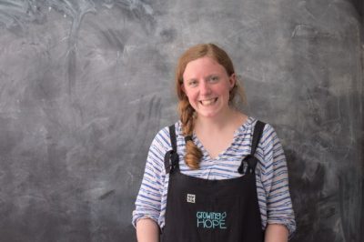 Dr Naomi Graham, wearing overalls with 'Growing Hope' written on the front, stands smiling in front of a recently scrubbed chalk blackboard